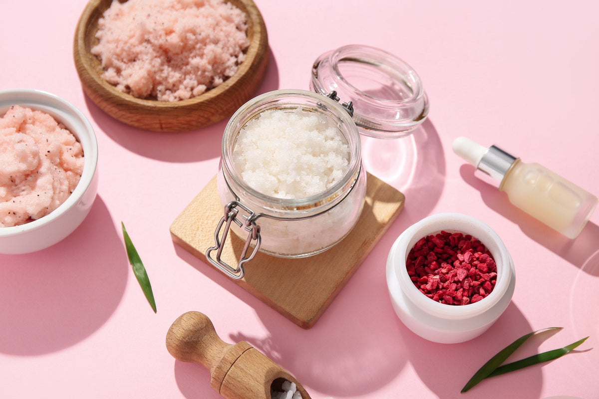 Jars of body scrub with an oil bottle and freeze-dried raspberries on a pink background