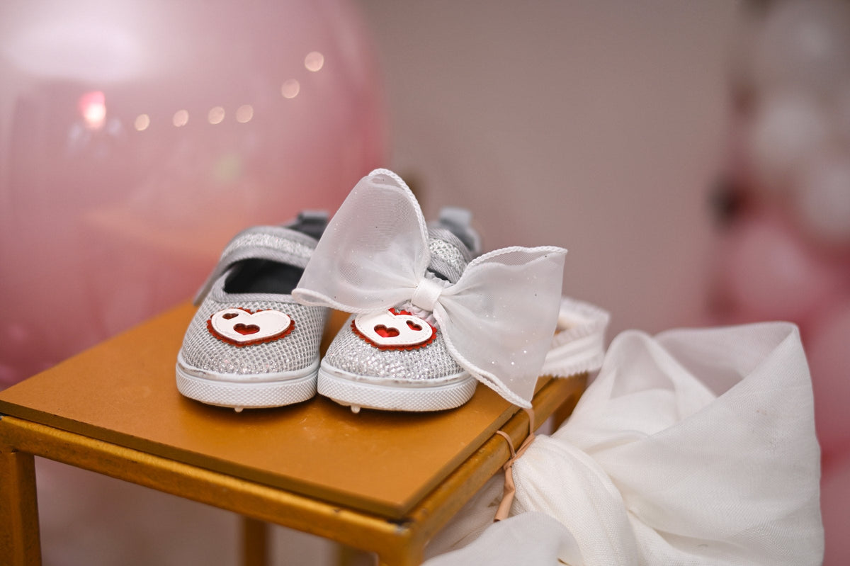 Small white baby shoes with bows on a wooden surface, with a pink balloon behind